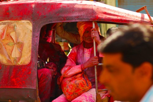 A three wheeled tuk-tuk (auto rickshaw) covered in red gulaal ferrying people within the city.