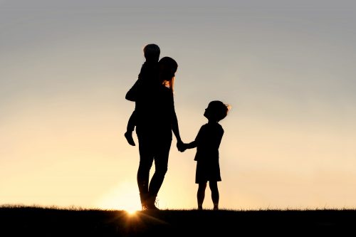 Silhouette of a young mother lovingly holding hands with her happy little child while holding his baby brother outside in front of a sunset in the sky.