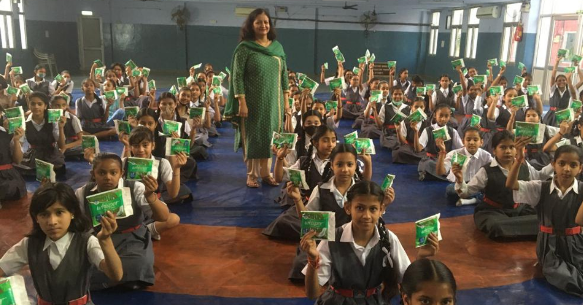 a school teacher in a green suit stands while girls in uniform sitting in rows hold up packets of sanitary napkins during a period workshop