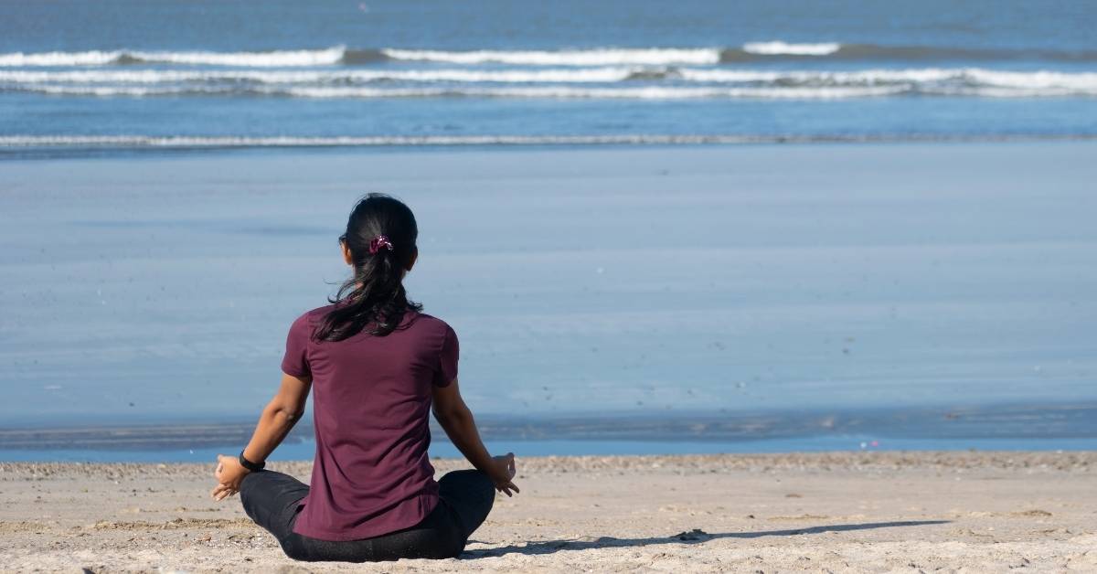 a woman dressed in yoga wear meditates on the beach with her back to the camera 