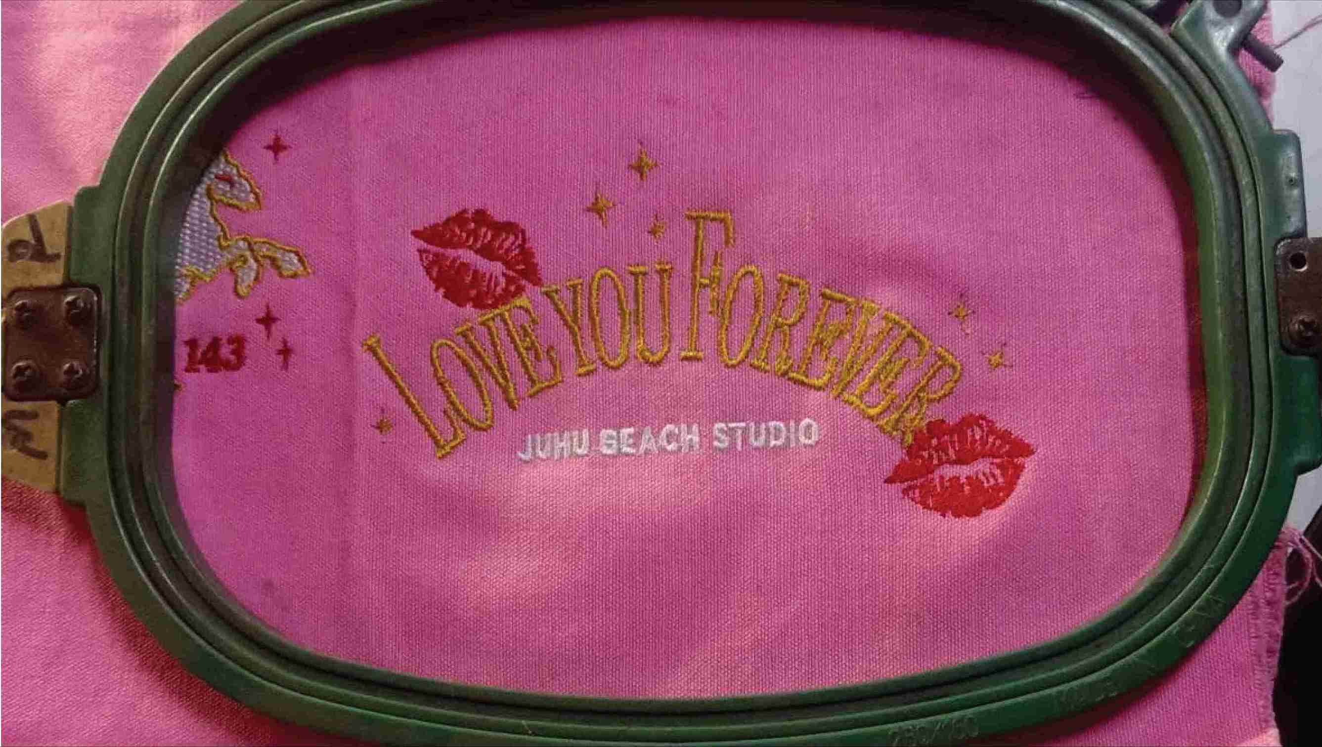 The phrase "Love You Forever"  beautifully embroidered in gold on a bright pink fabric, followed by the words "Juhu Beach Studio."