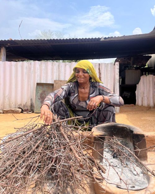 A woman clad in traditional banjara clothing, preparing her mud stove with wood fuel. 